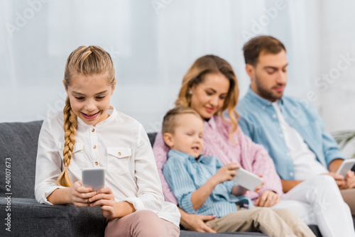 cute girl sitting on sofa and using smartphone with family at background in living room