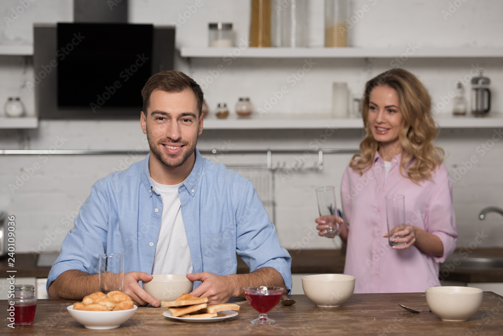 smiling man sitting at table and looking at camera with pretty wife holding glasses at background smiling man sitting at table and looking at camera with pretty wife holding glasses at background