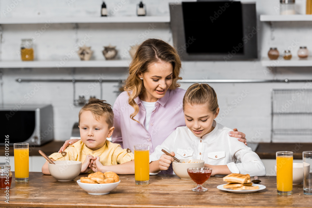 pretty woman embracing cute children eating oatmeal at kitchen table pretty woman embracing cute children eating oatmeal at kitchen table