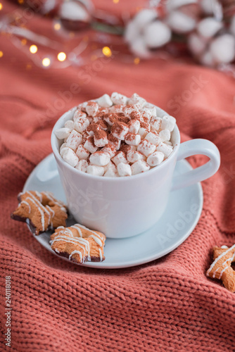 Coffee cup with marshmallow and stars cookies on warm knitted coral blanket, christmas lights and cotton flowers.