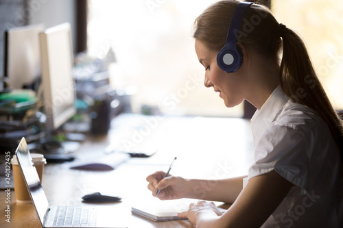 Focused woman wearing headphones write notes watch webinar study online with skype teacher, young female student learning computer course with laptop listening lecture, interpreter translating class photo