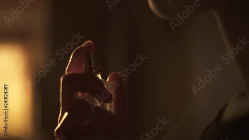 Young woman squirting perfume on her neck. In a room orange sunset light. Splashes fly slowly in the sunlight.
