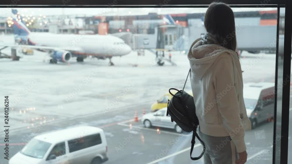 Backside view girl with backpack looks out window at plane and waves her hand. Backside view girl with backpack looks out window at plane and waves her hand.