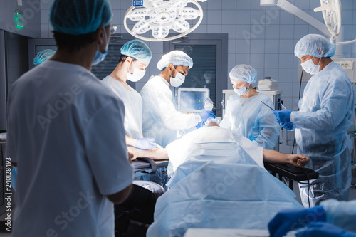 Serious skillful male surgeon standing in the operation theater and taking an instrument while the other team members assist him.