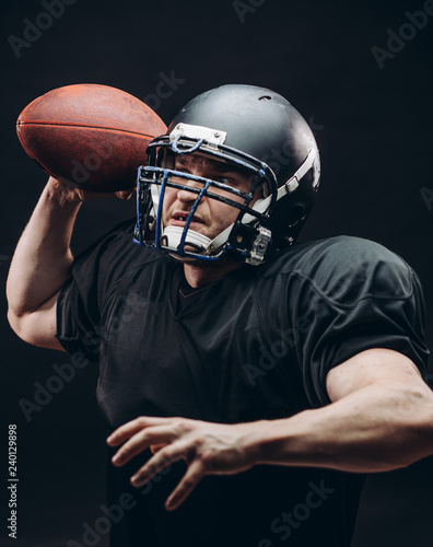 Isolated motion shot of American Football Quarterback player in professional protective black uniform and helmet passing a ball over black background.