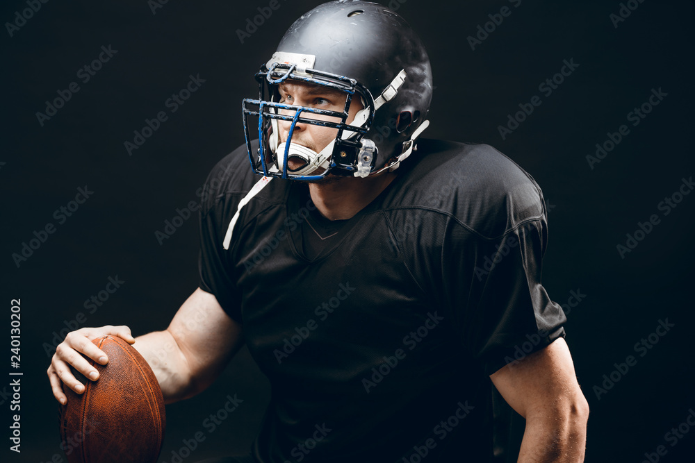Caucasian athlete in american football player uniform and black helmet holding oval ball posing over black background