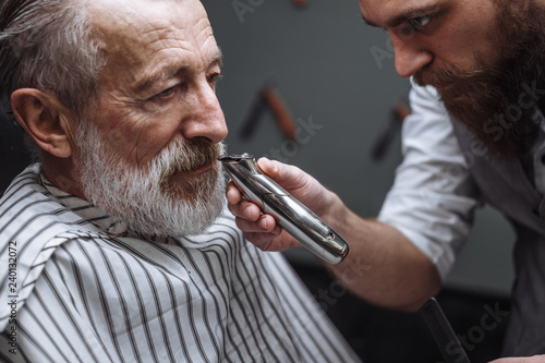 Close up of a hairdresser s work for a handsome respectable old-aged bearded man at the barber shop. He is doing styling with the electric shaver. photo