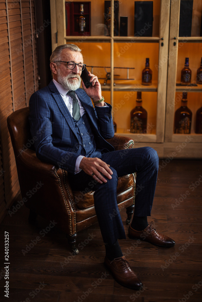 Experienced old private physician in spectacles and chic classy elegant suit talking on smartphone with his patient while resting in the big soft arm-chair of moder lounge restaurant or pub at night.