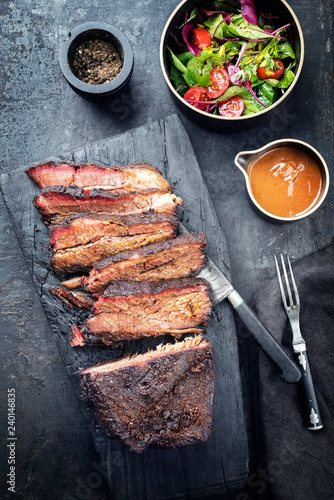 Traditional smoked barbecue wagyu beef brisket offered as top view on an old cutting board with Louisiana sauce and salad with tomatoes