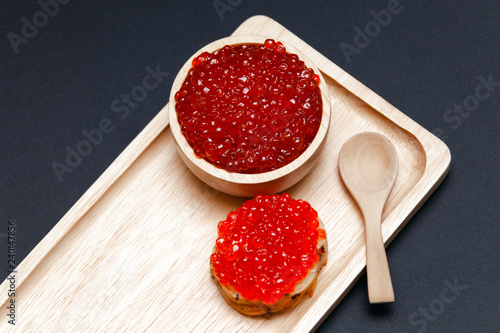 Closeup macro small red salmon caviar canape sandwich on white bread, saucer with slide, little spoon on rectangular wooden plate on black background. Concept gourmet snack seafood appetizer photo