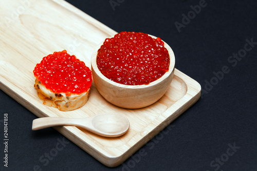 Closeup macro small red salmon caviar canape sandwich on white bread, saucer with slide, little spoon on rectangular wooden plate on black background. Concept gourmet snack seafood appetizer photo