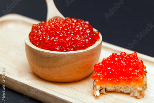 Closeup macro saucer with red salmon caviar with slide, bitten sandwich and little spoon on rectangular wooden plate on black background. Concept gourmet snack healthy seafood appetizer photo