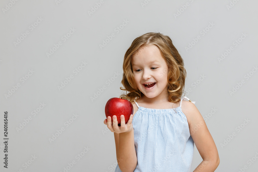 Portrait of a happy smiling little blonde girl on a gray background. The child is looking and ready to eat red apple Portrait of a happy smiling little blonde girl on a gray background. The child is looking and ready to eat red apple
