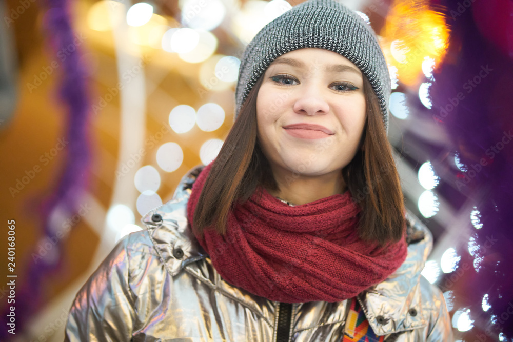 portrait of a smiling girl pleased. girl in a winter shiny jacket a red scarf and hat. on the background of Christmas lights. bokeh Garlands. portrait of a smiling girl pleased. girl in a winter shiny jacket a red scarf and hat. on the background of Christmas lights. bokeh Garlands.
