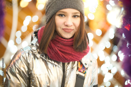 portrait of a smiling girl girl in a winter shiny jacket a red scarf and hat. on the background of Christmas lights. bokeh Garlands. © Alex