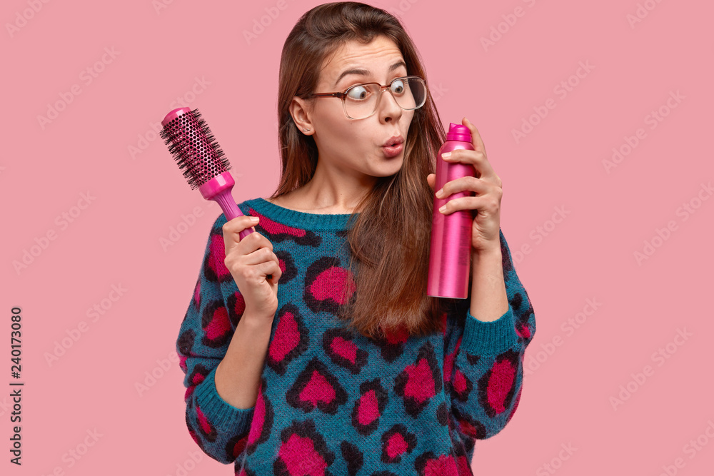 Attractive female wears big square glasses, stares with stupefaction at hairspray, holds hairbrush, wants to make coiffure for special occasion, models over pink background, going to fixate hair Attractive female wears big square glasses, stares with stupefaction at hairspray, holds hairbrush, wants to make coiffure for special occasion, models over pink background, going to fixate hair
