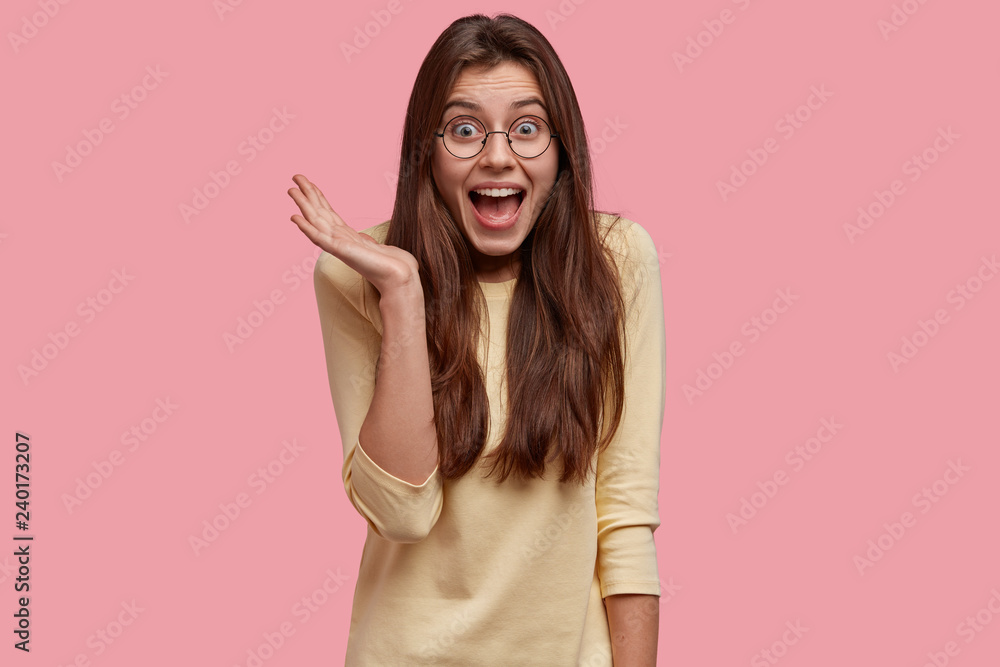 Overjoyed pretty lady keeps hand raised, exclaims with happiness, has long hair, dressed in casual outfit, isolated over pink background, expresses positive emotions. Wow, thats just amazing Overjoyed pretty lady keeps hand raised, exclaims with happiness, has long hair, dressed in casual outfit, isolated over pink background, expresses positive emotions. Wow, thats just amazing