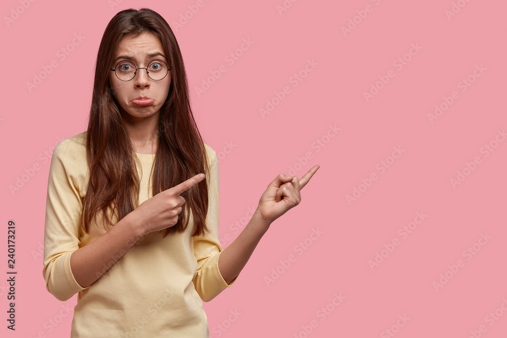 Offensive dejected lady purses lower lip, shows direction somewhere, indicates with both index fingers at free space, has gloomy facial expression, isolated on pink background. Promotion concept Offensive dejected lady purses lower lip, shows direction somewhere, indicates with both index fingers at free space, has gloomy facial expression, isolated on pink background. Promotion concept