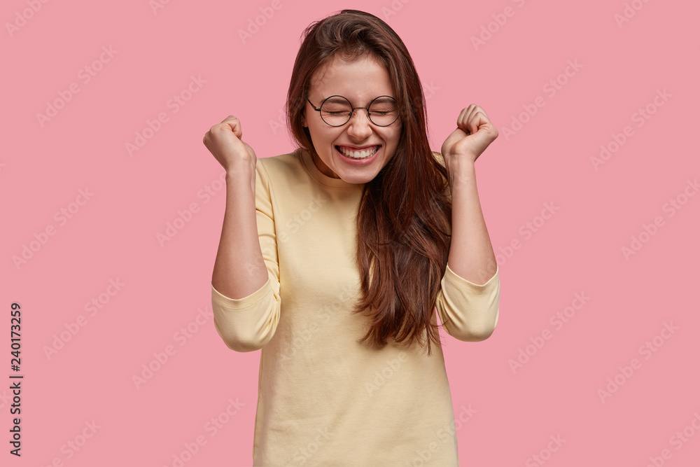 Overjoyed woman raises clenched fists in cheer, enjoys success and triumph, wears spectacles and casual outfit, models over pink background, celebrates winning competition, isolated on pink wall Overjoyed woman raises clenched fists in cheer, enjoys success and triumph, wears spectacles and casual outfit, models over pink background, celebrates winning competition, isolated on pink wall