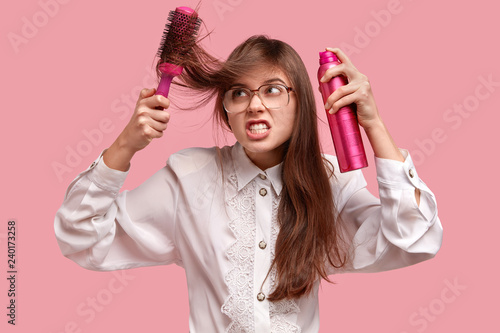 Irritated young lady makes hairstyle with hairspray and hairbrush, has ratty hair, wears big spectacles and white old fashionable blouse, models over pink studio wall. Beauty and care concept photo