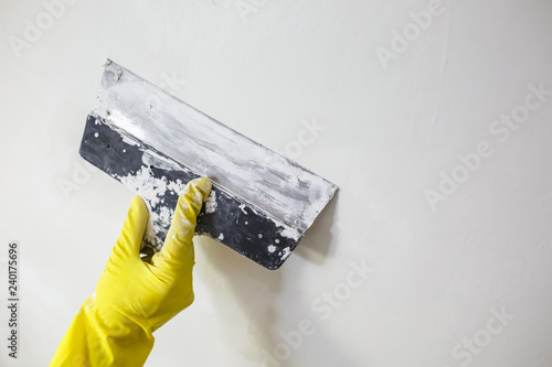 worker's hand in yellow gloves holding putty knife patching a hole with spatula with plaster or putty in white wall. Renovation and repair process, remodeling interior of room at apartment building photo