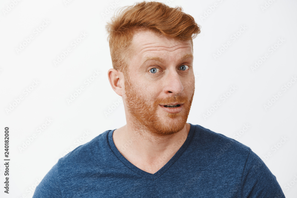 Headshot of intrigued good-looking ginger male with bristle in blue t-shirt turning at camera with impressed and curious expression, listening to interesting story carefully over gray background Headshot of intrigued good-looking ginger male with bristle in blue t-shirt turning at camera with impressed and curious expression, listening to interesting story carefully over gray background