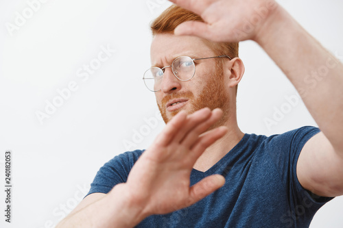 Take this ugly thing away from me. Displeased intense redhead mature guy in glasses and blue t-shirt, turning away and protecting face with raised hands, seeing something disgusting over grey wall