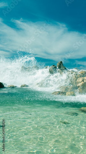 Ocean waves break over a rock wall sending spray into the air. Bright blue sky and clear turquoise water of an island paradise coastal scene.