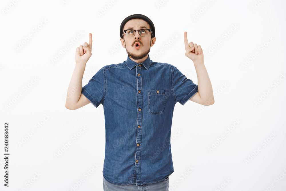 Guy being impressed of seeing amazing copy space. Stylish young european male with beard and moustache in hat and glasses folding lips saying wow pointing and staring up excited over white wall Guy being impressed of seeing amazing copy space. Stylish young european male with beard and moustache in hat and glasses folding lips saying wow pointing and staring up excited over white wall