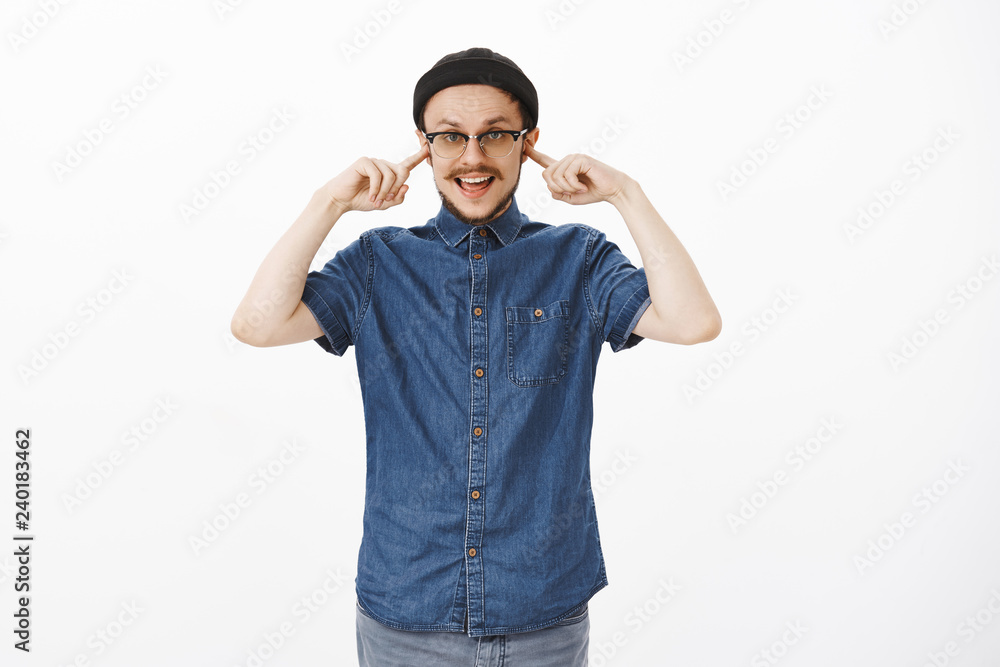 Fototapeta premium Cannot hear your boring talks. Carefree indifferent handsome stylish male with moustache in black beanie and glasses covering ears with index finger not listen girlfriend yell for him comming late