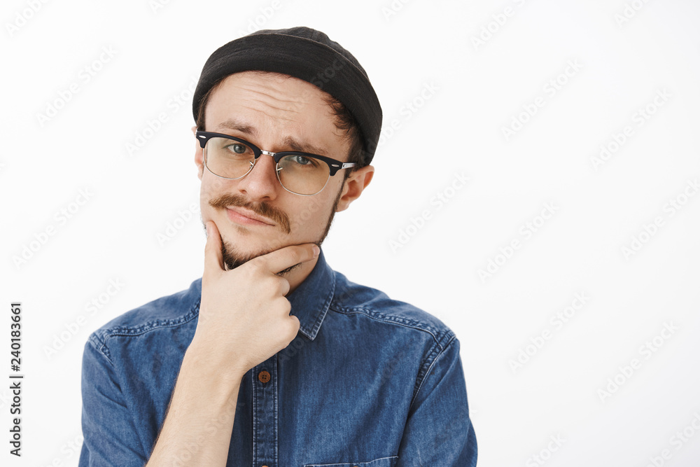 Close-up shot of doubtful smart suspicious handsome young guy with beard and moustache in black beanie and glasses touching chin and smirking squinting at camera feeling uncertain and unimpressed Close-up shot of doubtful smart suspicious handsome young guy with beard and moustache in black beanie and glasses touching chin and smirking squinting at camera feeling uncertain and unimpressed