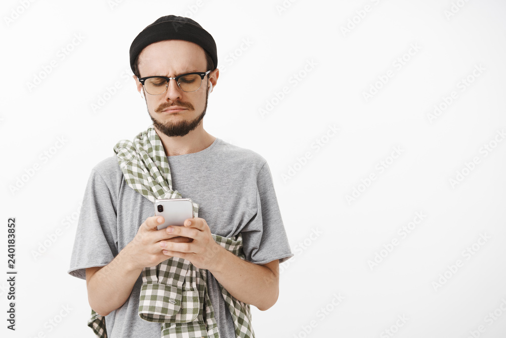 Gloomy unhappy and displeased handsome young bearded guy in glasses black beanie and checked shirt crossed on chest frowning making sad smile while staring with regret at smartphone screen Gloomy unhappy and displeased handsome young bearded guy in glasses black beanie and checked shirt crossed on chest frowning making sad smile while staring with regret at smartphone screen