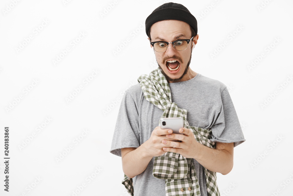 Waist-up shot of panicking shocked guy sending naked pick to mom by mistake shouting and staring shook at smartphone screen wearing wireless earbuds and black beanie over white wall Waist-up shot of panicking shocked guy sending naked pick to mom by mistake shouting and staring shook at smartphone screen wearing wireless earbuds and black beanie over white wall
