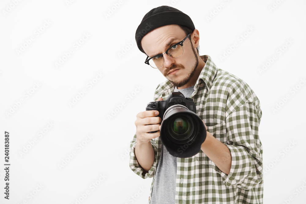 Obraz premium Waist-up shot of funny serious-looking male photographer in black beanie glasses and checked shirt pointing professional camera forward and gazing seriously to take picture during work