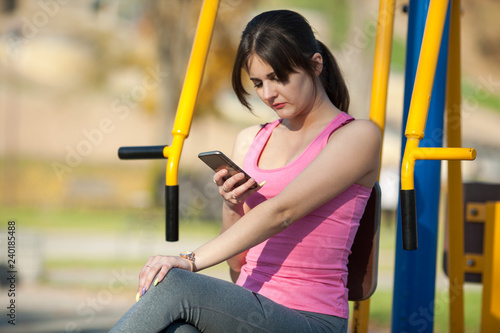 Young athletic girl in sunglasses is sitting on a sports simulator and surfing the internet in her smartphone on a street playground.