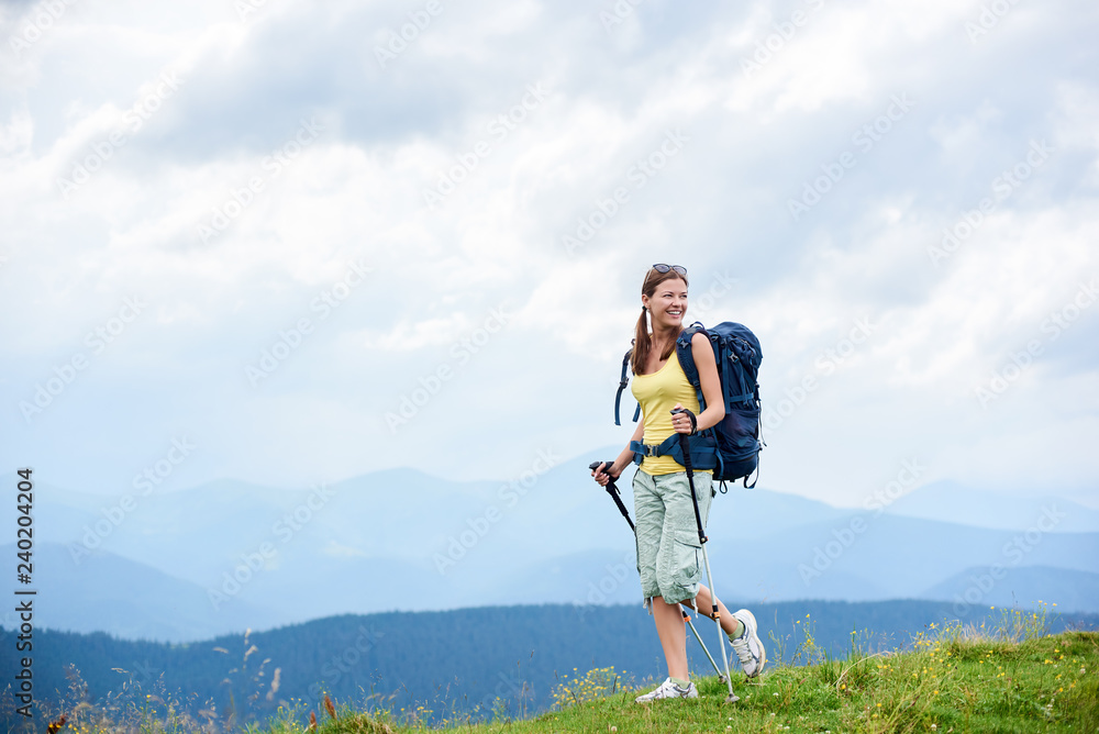 Attractive happy woman tourist hiking mountain trail, walking on grassy hill, wearing backpack, using trekking sticks, enjoying summer cloudy day. Outdoor activity, tourism concept