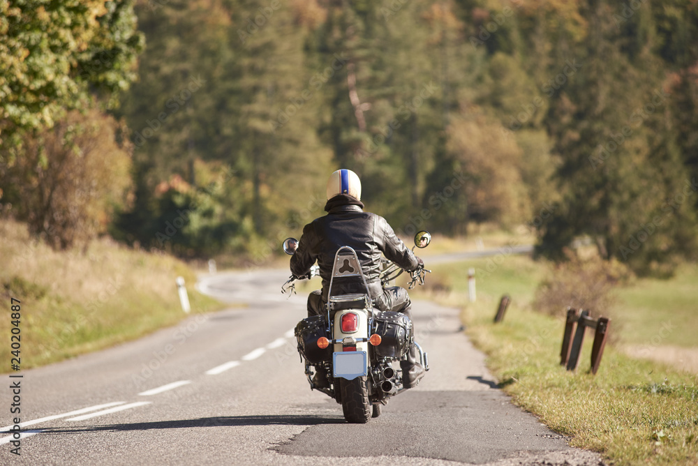 Back view of biker in black leather jacket and helmet riding motorcycle along twisty road on blurred background of dense green forest trees on bright sunny summer day. Active lifestyle and traveling.