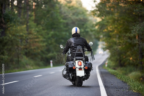 Back view of biker in black leather jacket and white helmet riding motorbike along hilly road between tall green trees. Active lifestyle, love to adventures concept.