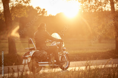 Side view of bearded motorcyclist riding modern powerful cruiser motorcycle along empty narrow country road at sunset on beautifull golden autumn landscape background.