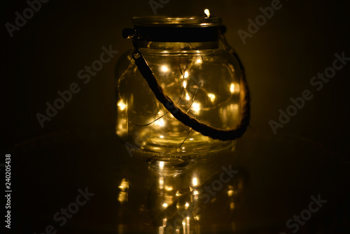 Decorative lights in glass jar at the dark room with mirror effect on dark background. Festive decorations .Warm white led lights