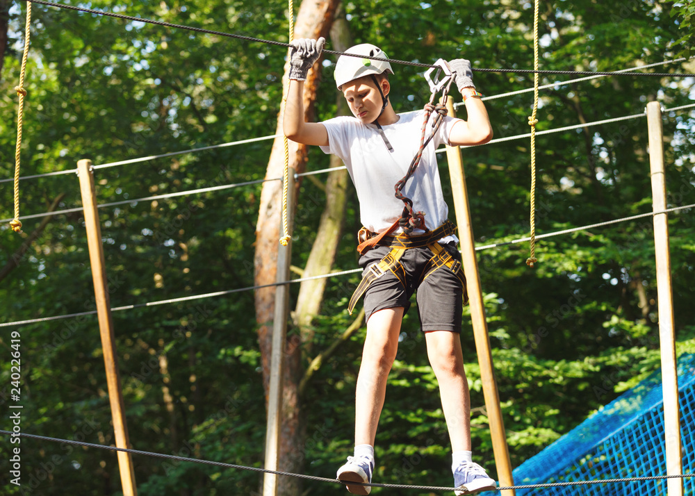 Cute boy enjoying activity in climbing adventure park at sunny summer day. Kid climbing in rope playground structure. Safe climbing with helmet insurance. Child in forest adventure park, extreme sport