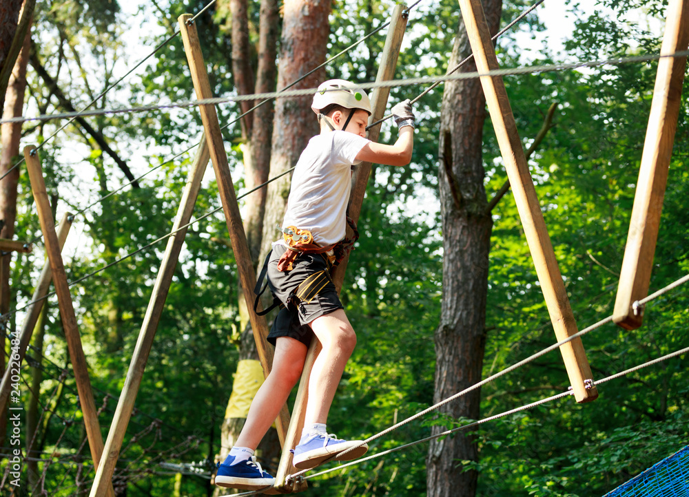 Cute boy enjoying activity in climbing adventure park at sunny summer day. Kid climbing in rope playground structure. Safe climbing with helmet insurance. Child in forest adventure park, extreme sport