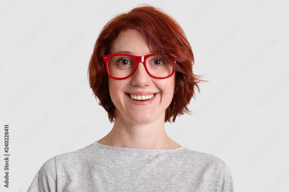 Close up shot of smiling glad woman with short hairstyle, wears red rimed spectacles, dressed casually, isolated over white background, expresses positive feelings. People and beauty concept.