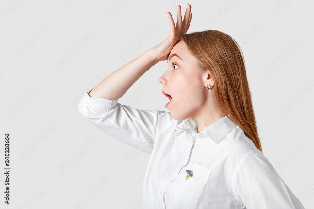 Fototapeta premium Surprised forgetful beautiful woman remembers something too late, holds hand on forehead, opens mouth widely, stands sideways against white background, dressed in formal shirt. Memory concept
