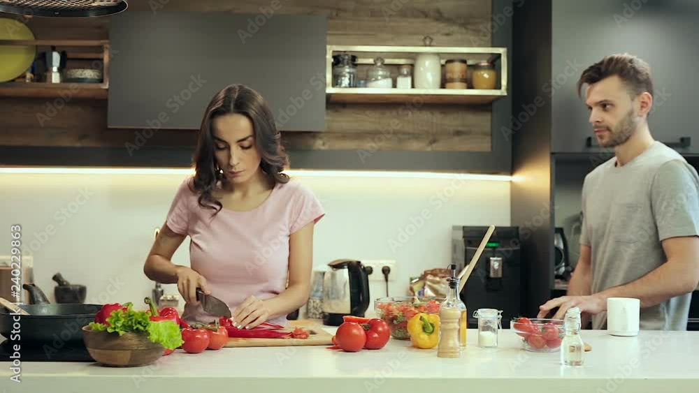 Young couple cooking and having fun. Man in good mood fooling around and taking peace of pepper, while woman preparing salad.