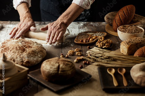Hands kneading raw dough on table. Top view on baker workplace, working with pastry, all surface on flowered table is occupied with loaves of bread and ingredients. Culinary, cooking, bakery concept