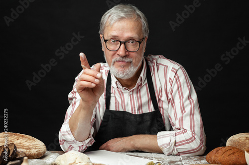 Portrait of senior grey-bearded baker, small businee owner, demonstrates his products in bakery, standing with different kinds of fresh bread on wooden table. photo