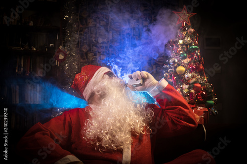 Santa Clause vaping electronic cigarette dressed as traditional Santa on a dark toned background with vape clouds. Selective focus.