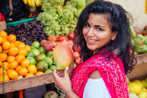 Portrait of beautiful traditional Indian woman in red sari dress choosing mango fruits in a street market in india.Happy girl shopper in a supermarket fruit shop.exotic sale Kerala Goa photo