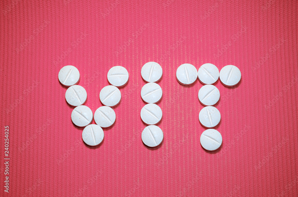 Lots of white smooth convex tablets with dividing strip on pink striped background, in the form of word VIT, underlined, vitamins, top view Lots of white smooth convex tablets with dividing strip on pink striped background, in the form of word VIT, underlined, vitamins, top view